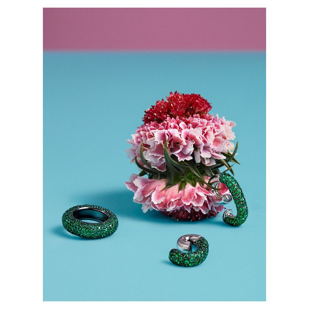 Three rings on blue background with carnation flowers in pink and red on top. Emerald green sparkly design on two rings.