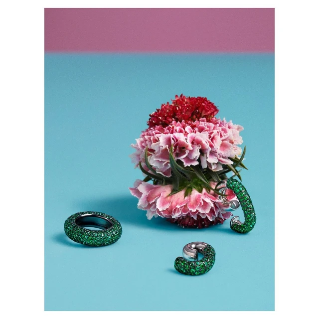 Three rings on blue background with carnation flowers in pink and red on top. Emerald green sparkly design on two rings.