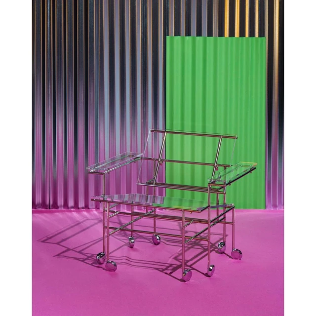 Modern transparent chair with metallic frame and wheels on a pink floor against a wavy metallic and green backdrop, studio lighting.