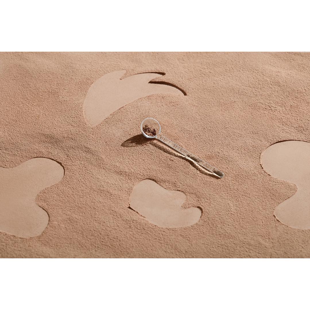 Small spoon with seeds on beige textured background with irregular cutout shapes creating a minimalist still life scene.