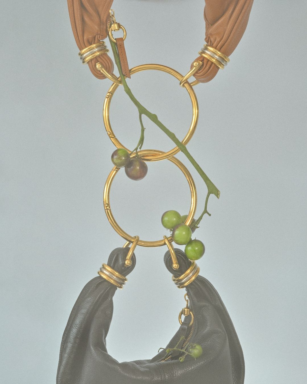 Fashion handbag with golden rings, suspended and decorated with green and brown berries against a light blue background in studio lighting.