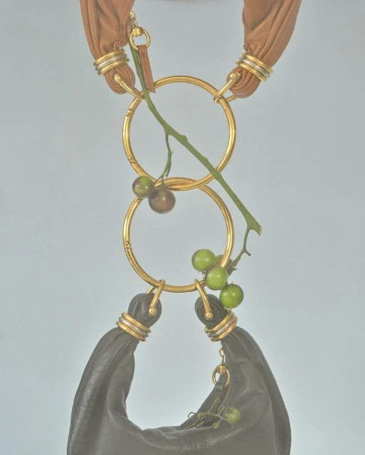Fashion handbag with golden rings, suspended and decorated with green and brown berries against a light blue background in studio lighting.