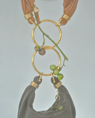 Fashion handbag with golden rings, suspended and decorated with green and brown berries against a light blue background in studio lighting.
