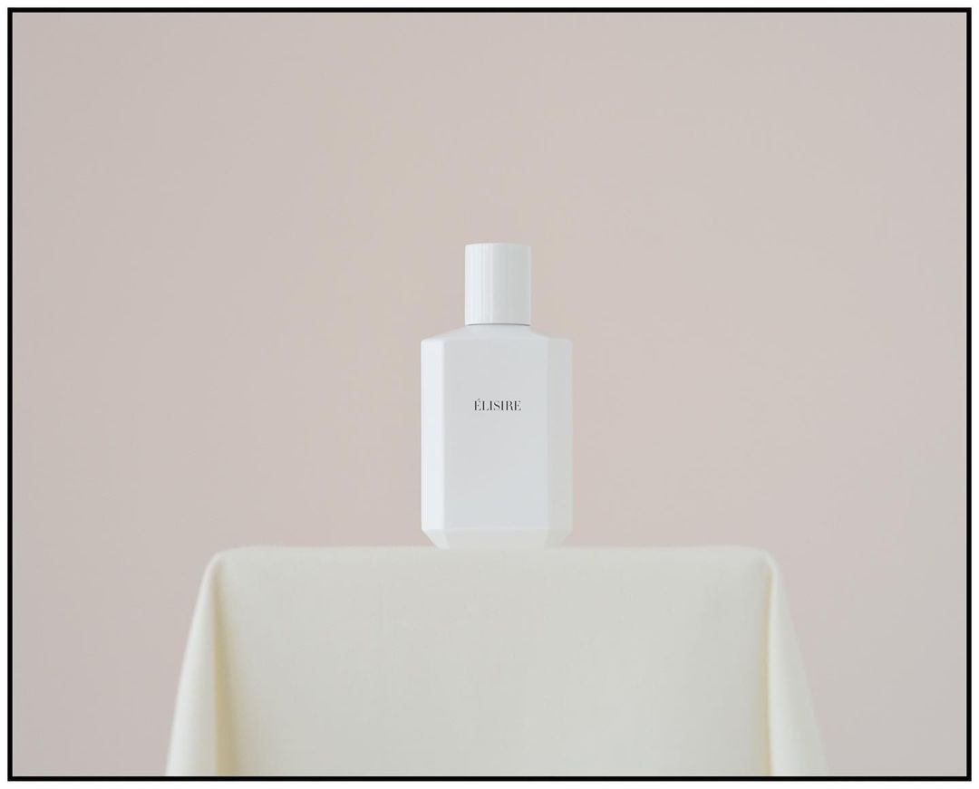 Elegant studio shot of a white 'ELISIRE' perfume bottle on a draped table against a soft cream background; minimalist aesthetic.
