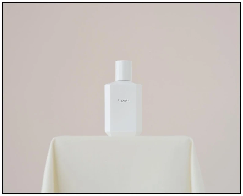 Elegant studio shot of a white 'ELISIRE' perfume bottle on a draped table against a soft cream background; minimalist aesthetic.