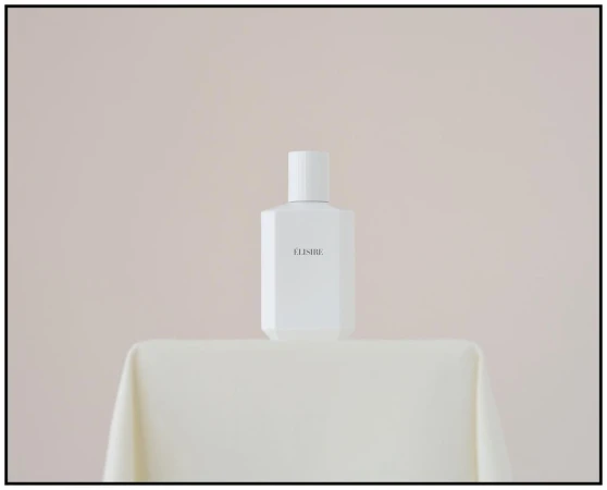 Elegant studio shot of a white 'ELISIRE' perfume bottle on a draped table against a soft cream background; minimalist aesthetic.