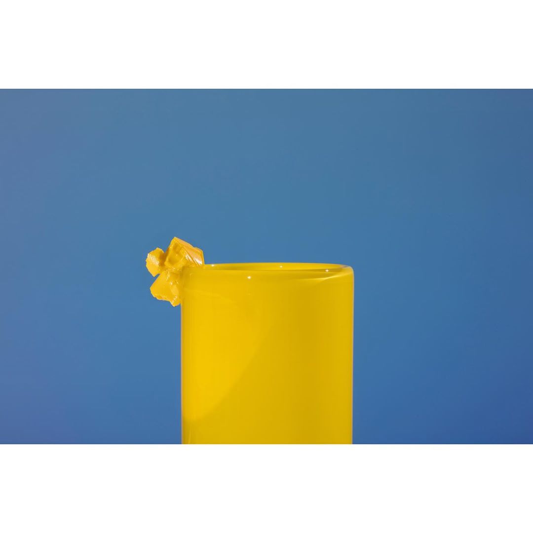 Close-up studio shot of a cylindrical yellow vase with a decorative yellow flourish against a bright blue seamless backdrop.