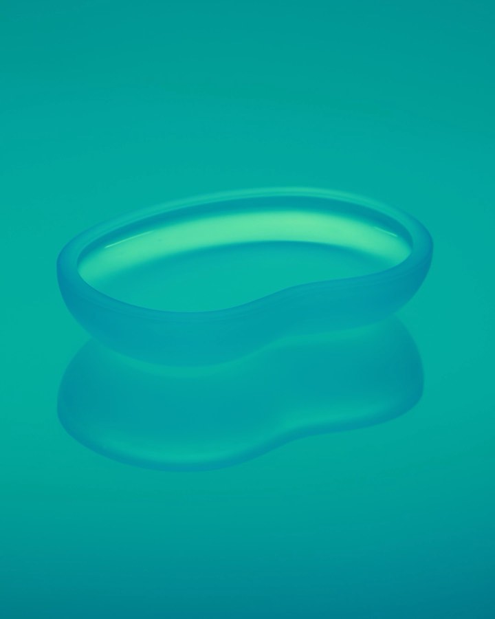 Translucent blue glass dish with a smooth, curved design on a matching reflective blue background. Minimalist studio product shot.