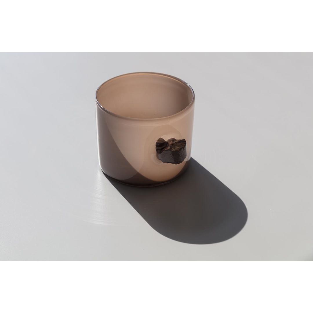 Beige glass cup with a solid brown rock-like handle on a white background with a shadow suggesting natural light in a studio.