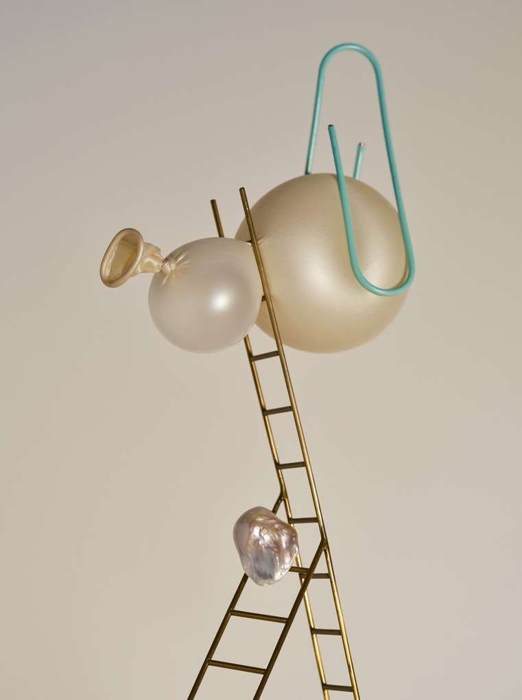 Surreal still life featuring translucent balloons, a mini-ladder, a baroque pearl, and a blue paperclip on a cream-colored backdrop.