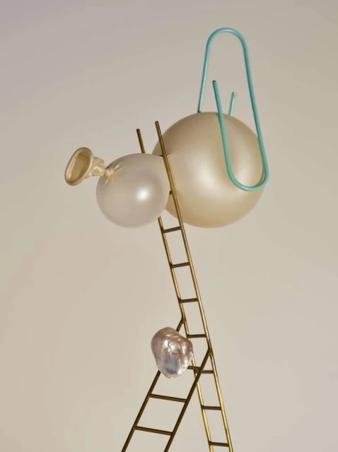 Surreal still life featuring translucent balloons, a mini-ladder, a baroque pearl, and a blue paperclip on a cream-colored backdrop.