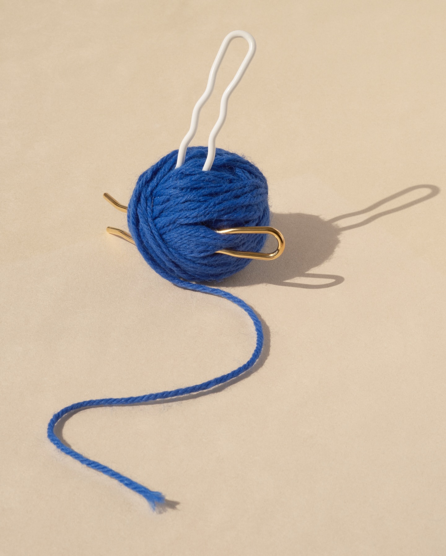Close-up of a blue ball of yarn with white and gold yarn needles inserted into it against a tan background in soft lighting.