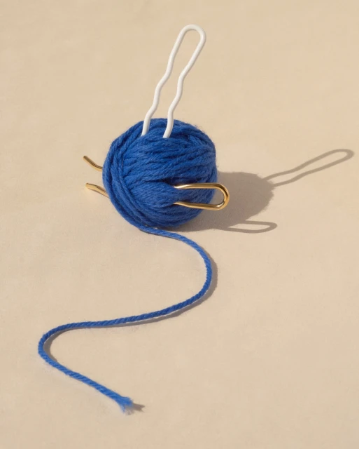 Close-up of a blue ball of yarn with white and gold yarn needles inserted into it against a tan background in soft lighting.
