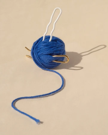Close-up of a blue ball of yarn with white and gold yarn needles inserted into it against a tan background in soft lighting.