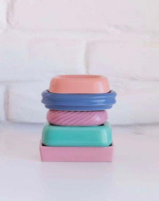 Stack of colorful ceramic dishes and bowls of pink, blue, and green hues arranged on a white surface against a textured white background.