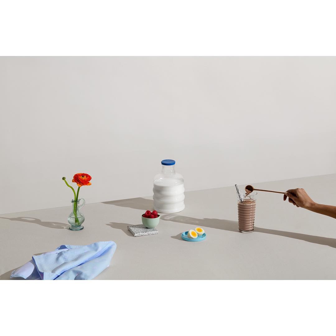 Still life with a chocolate milkshake, milk bottle, flower in a vase, raspberries, sliced boiled eggs, and a light blue cloth on a table.