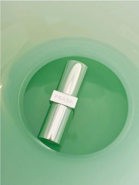 Close-up studio shot of a silver lipstick tube with the brand Prada on it, set against a green background. Beauty and fashion photography.