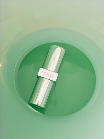 Close-up studio shot of a silver lipstick tube with the brand Prada on it, set against a green background. Beauty and fashion photography.