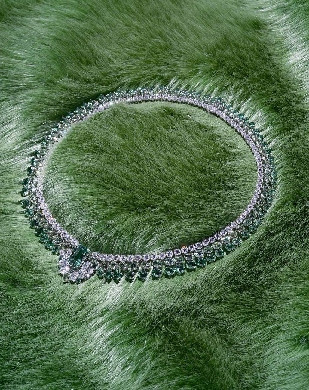 Diamond and emerald necklace displayed on a light green fur surface, showcasing fine jewelry and luxurious materials.