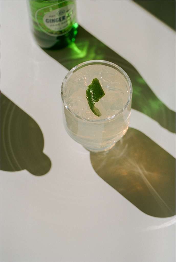 Overhead shot of a cocktail in a clear glass with lime peel garnish, set on a reflective white surface with shadows.