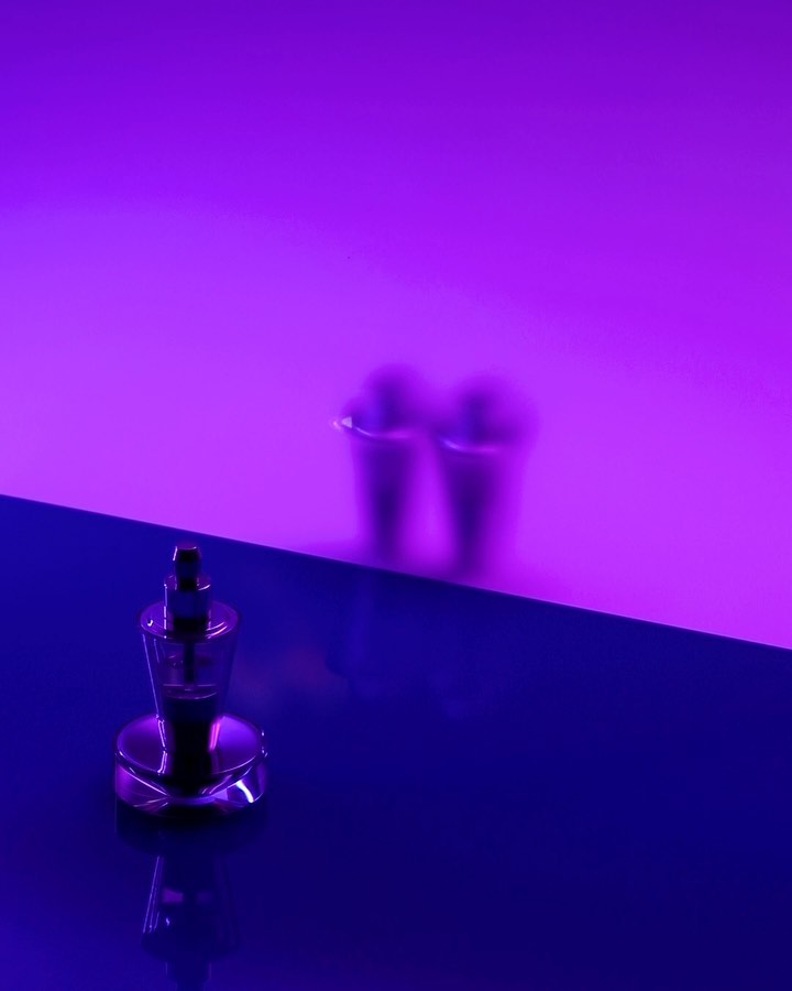 Perfume bottle with chrome accents on a blue surface, cups blurred in background, highlighted by purple and pink lighting, studio shot.