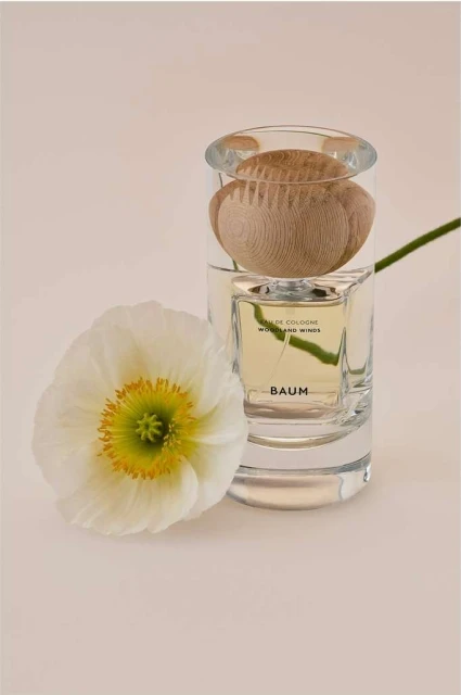 Close-up studio shot of a transparent perfume bottle with a wooden cap, next to a single white poppy flower against an off-white backdrop.