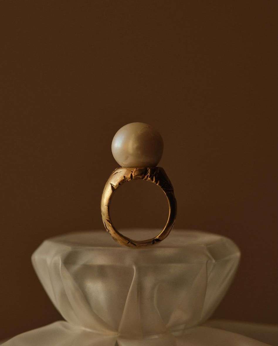 Close-up of a gold ring with an off-white pearl on top, placed on a frosted glass surface, and set against a dark brown background.