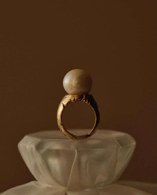 Close-up of a gold ring with an off-white pearl on top, placed on a frosted glass surface, and set against a dark brown background.