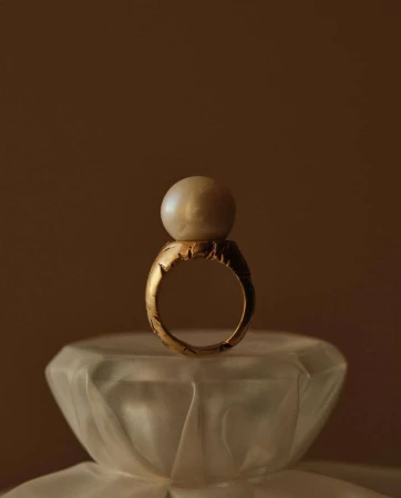 Close-up of a gold ring with an off-white pearl on top, placed on a frosted glass surface, and set against a dark brown background.
