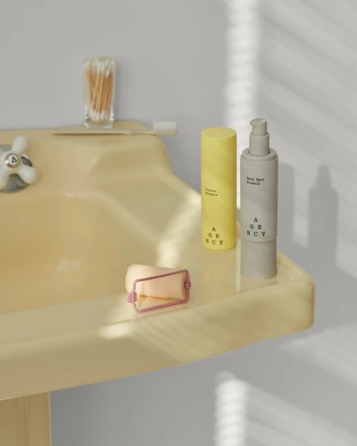 Agency skincare products arranged on a beige bathroom sink with a cotton swab holder and toothbrush against a neutral background.