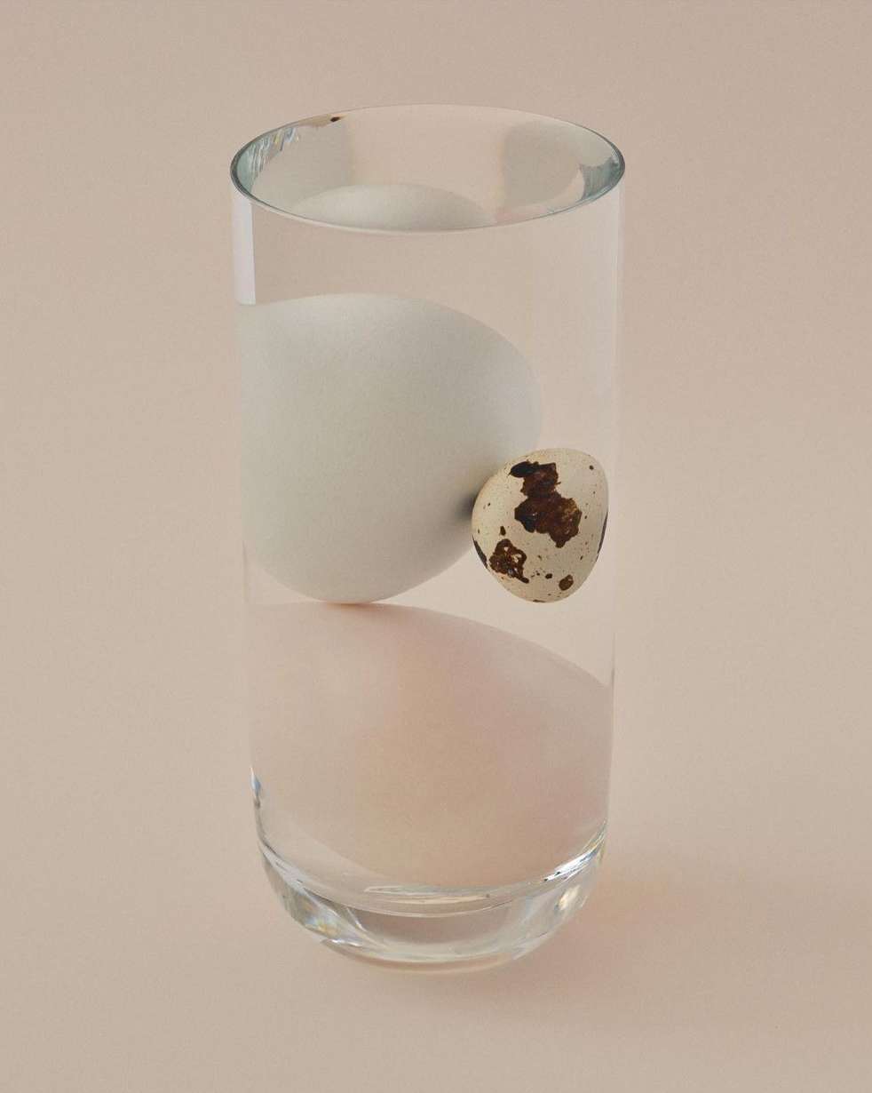 Three eggs—one large and white, one quail egg—inside a clear glass on a light tan backdrop. Food and beverage still life.