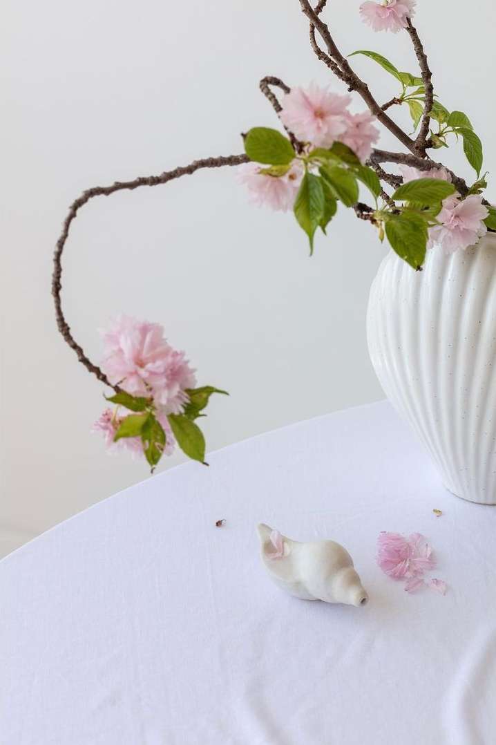 Elegant still life of pink blossoms arranged in a white vase on a white tablecloth with a small ceramic snail and petals.