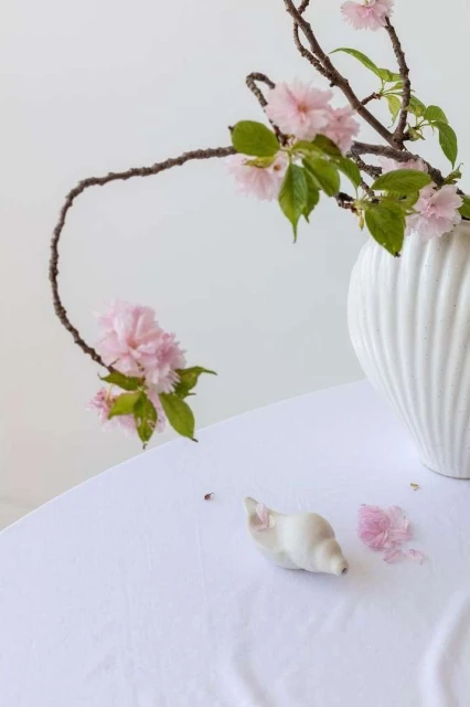 Elegant still life of pink blossoms arranged in a white vase on a white tablecloth with a small ceramic snail and petals.