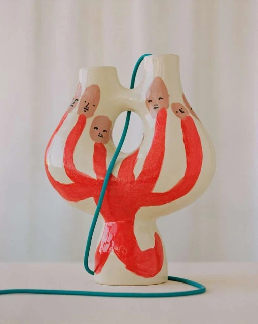 Handcrafted ceramic vase featuring painted faces and red painted bodies. Displayed on a white surface with a blue cord accent.