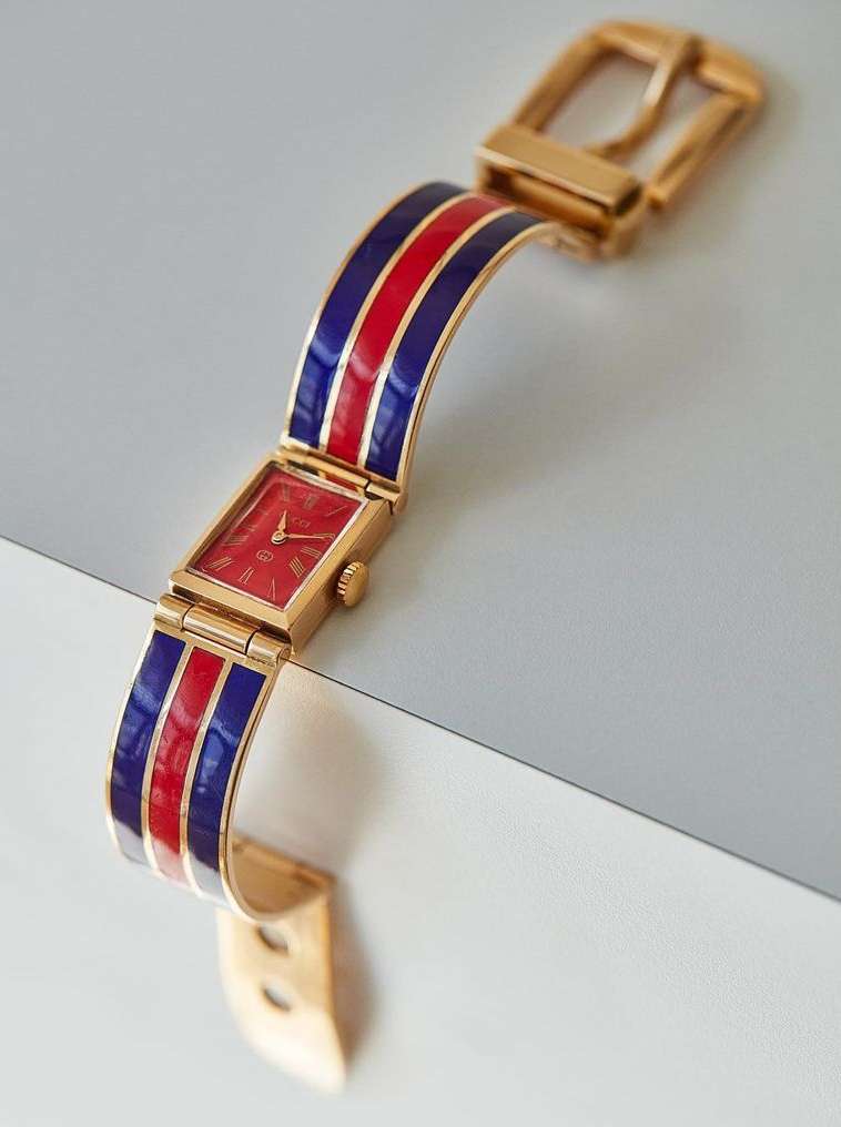 Gold wristwatch with a red watch face and a band featuring red and navy blue stripes, set against a white surface.
