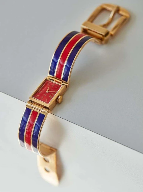 Gold wristwatch with a red watch face and a band featuring red and navy blue stripes, set against a white surface.