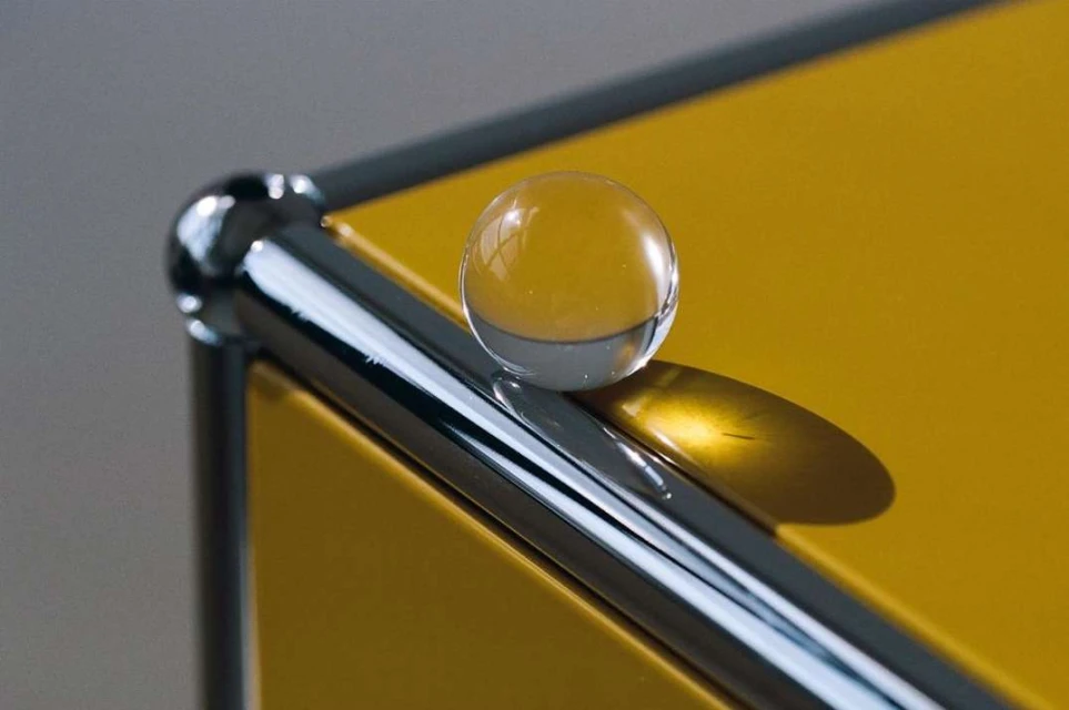 Close-up of a glass marble resting on a yellow and silver metal shelf. The sphere casts a shadow on the bright yellow surface.