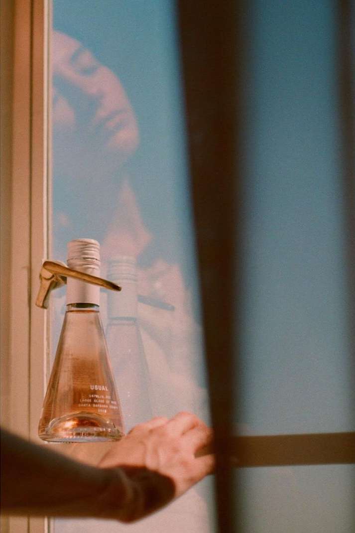 Rosé wine bottle on a window sill with a hand reaching to open the window. Golden light casting a shadow.