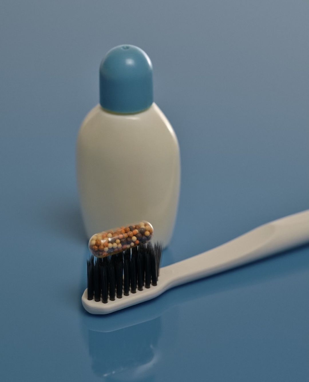 Toothbrush with black bristles, clear capsule filled with tiny colorful beads on top, white and blue bottle on a blue backdrop.