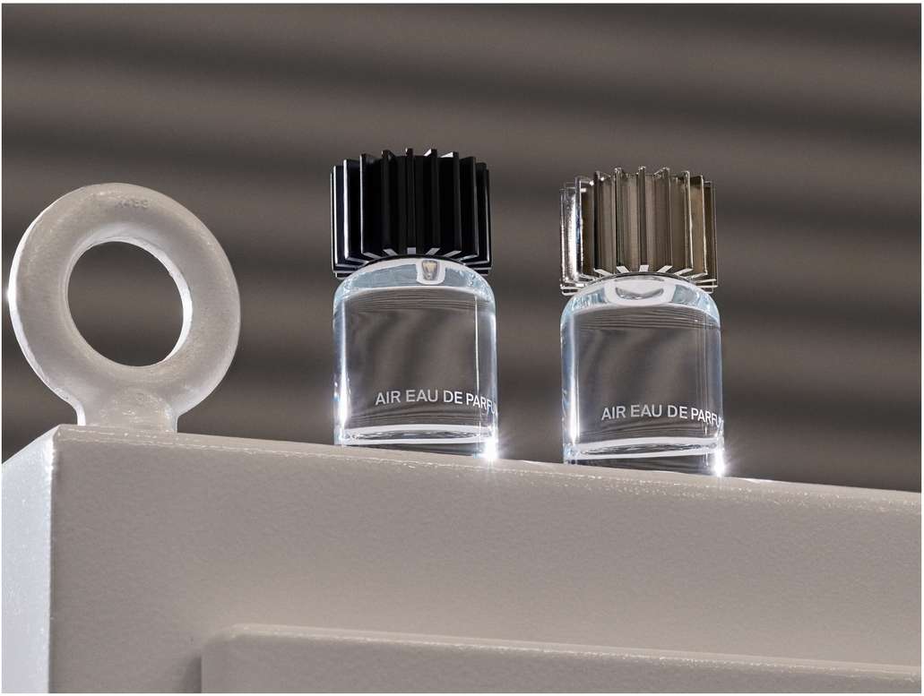 Two clear glass perfume bottles with black and silver caps sit on a white platform, against a textured gray background.