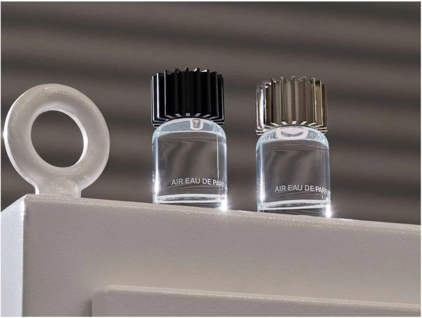 Two clear glass perfume bottles with black and silver caps sit on a white platform, against a textured gray background.