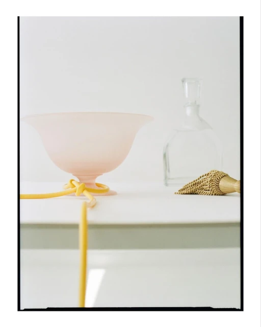 Still life of a pink glass bowl with a yellow ribbon, a clear glass bottle, and a tassel, set against a clean white background.