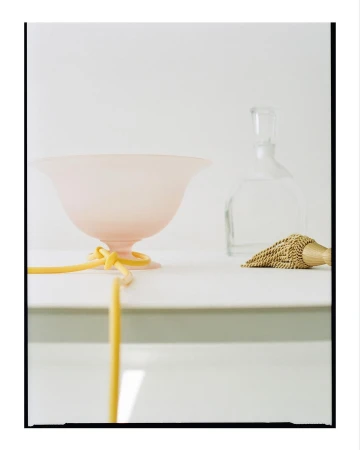 Still life of a pink glass bowl with a yellow ribbon, a clear glass bottle, and a tassel, set against a clean white background.