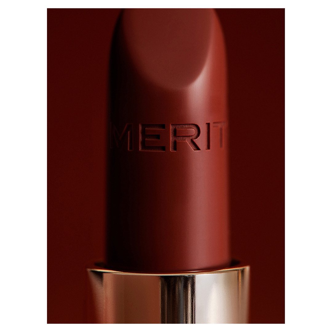 Close-up of a brown Merit lipstick with gold trim against a matching brown background, capturing its texture and simple packaging.