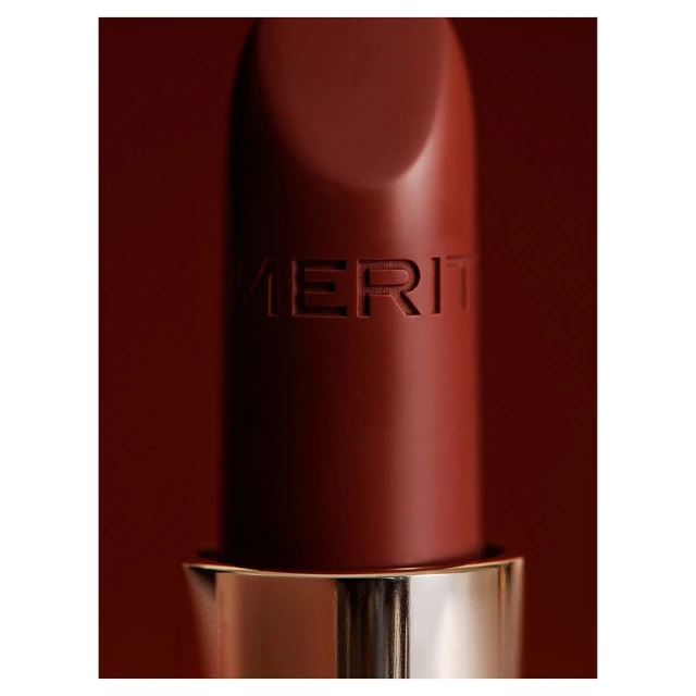 Close-up of a brown Merit lipstick with gold trim against a matching brown background, capturing its texture and simple packaging.