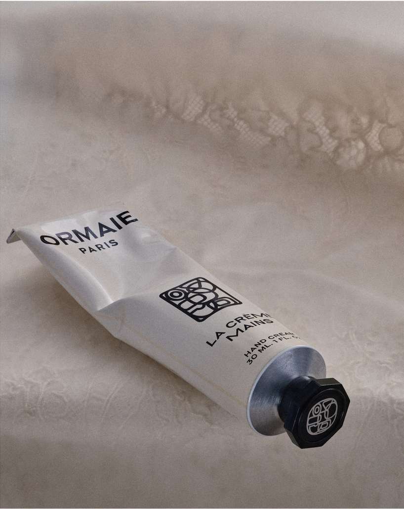 Close-up studio shot of a white Ormaie Paris hand cream tube with black cap set against a blurred lace fabric background.