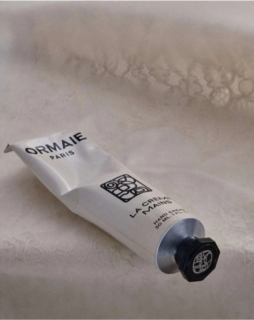 Close-up studio shot of a white Ormaie Paris hand cream tube with black cap set against a blurred lace fabric background.