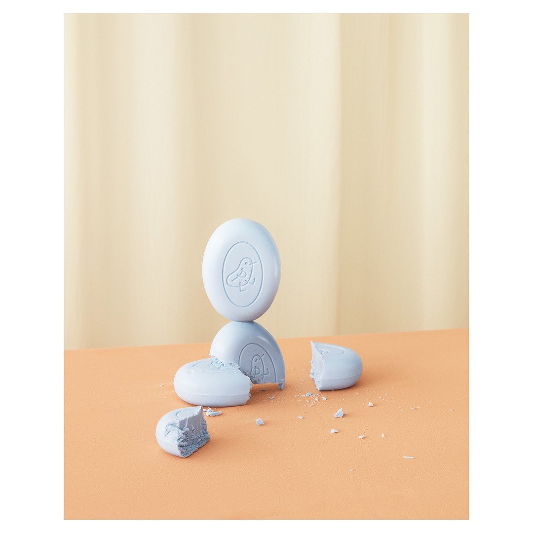 Still life shot of stacked oval-shaped soaps with a bird logo against a peach-colored surface and a cream-colored backdrop.