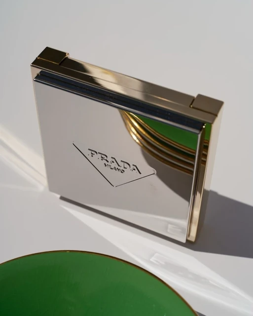 Elevated view of a silver Prada Milano compact with gold trim, alongside a green and gold saucer against a white background.