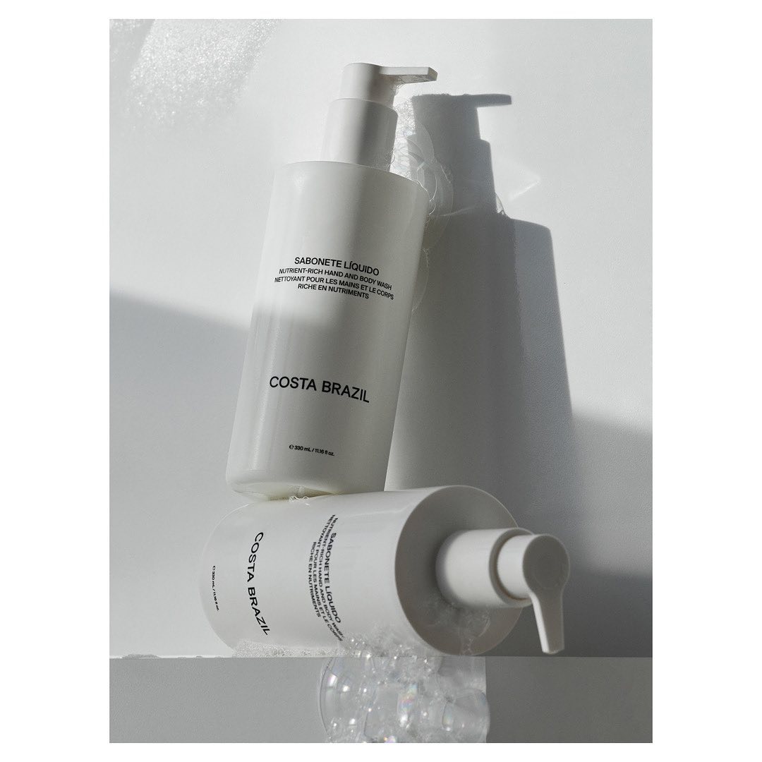 Two Costa Brazil Sabonete Liquido bottles with pump dispensers, one lying down and one standing, against a bright white background with bubbles.